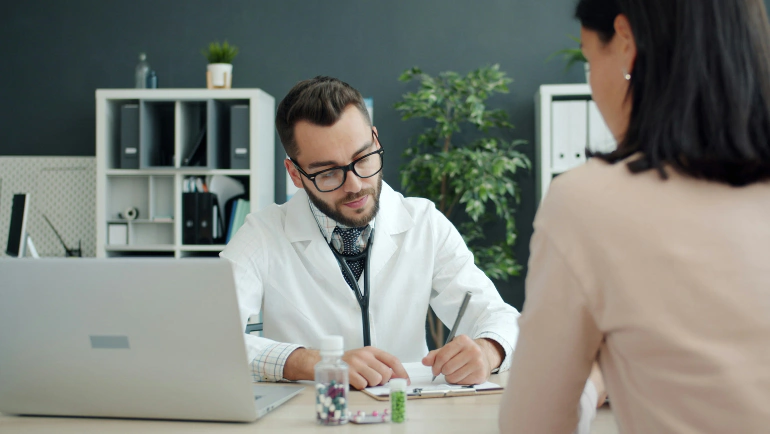 Doctor in a white coat writing notes while consulting with a patient at a desk with a laptop and medication bottles, illustrating recurring care programs and complex healthcare services.
