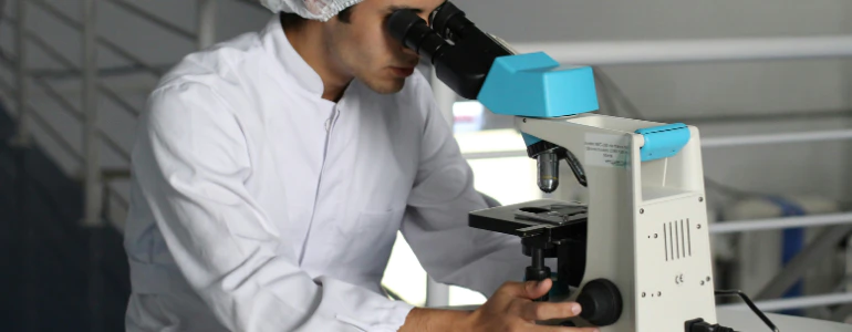 Lab technician in a white coat and hairnet looking through a microscope, illustrating payment processing for specialty healthcare businesses.